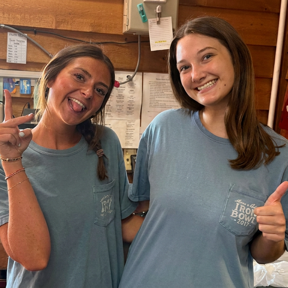 Two smiling restaurant workers in blue shirts pose inside a rustic wooden kitchen, giving a peace sign and thumbs-up.